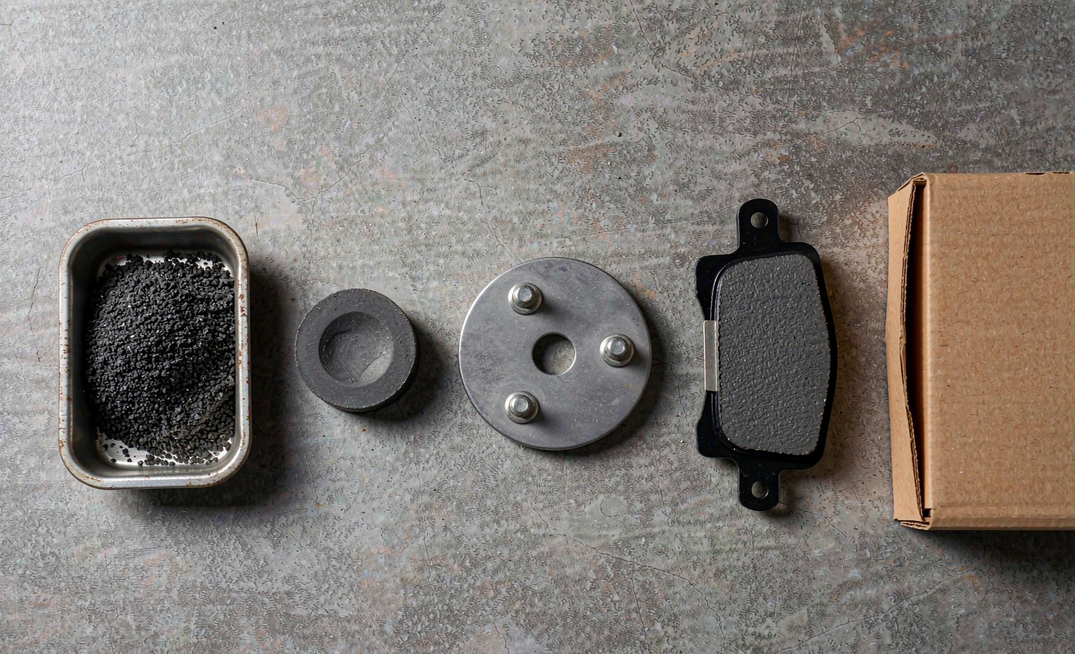 Brake pad cost stack shown as five production stages: friction compound granules, pressed puck, steel backing plate, finished pad and carton