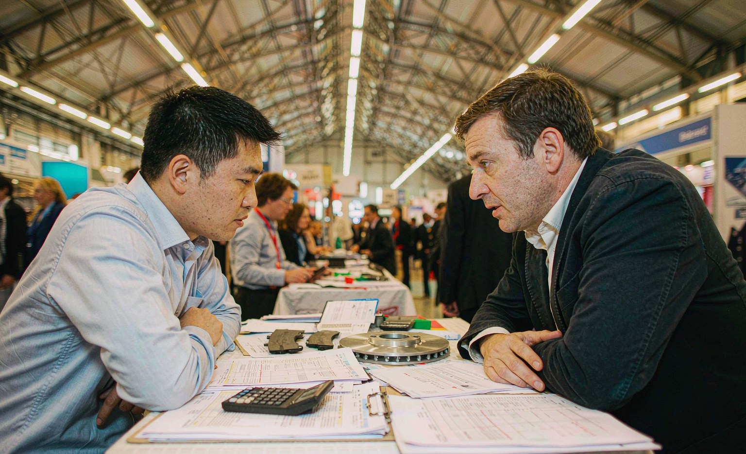 Buyer and supplier reviewing an auto parts quotation with brake pad and rotor samples at a trade fair table