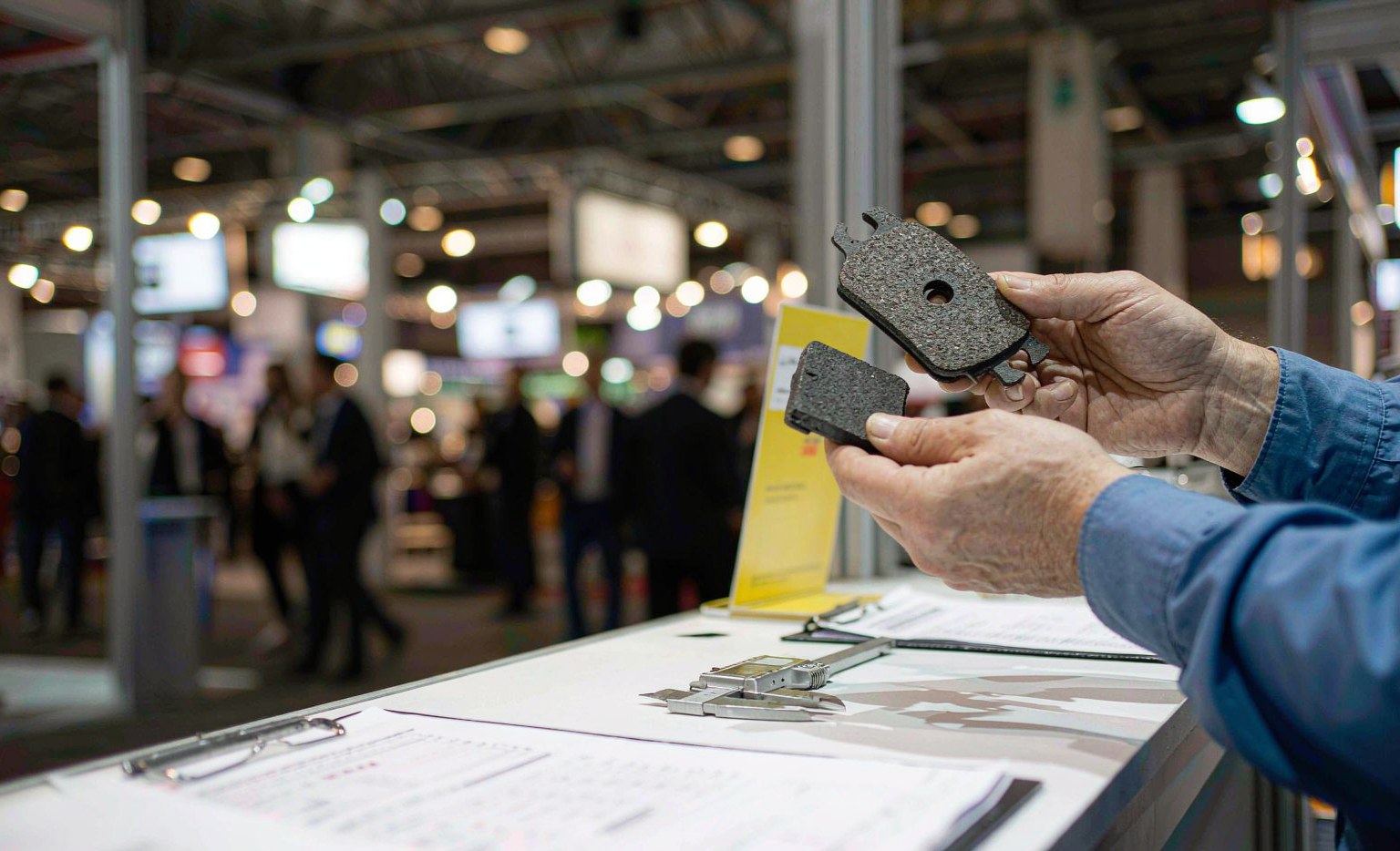 Two hands comparing brake pad samples on a trade show booth table with a digital caliper, blurred exhibition hall aisle in the background at Canton Fair auto parts sourcing