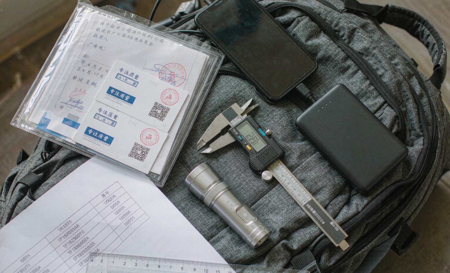 Flat-lay packing scene for an overseas trade fair trip with a Canton Fair buyer checklist: clear folder with invitation letter and business cards, smartphone, power bank, digital caliper, ruler, and an OE reference sheet laid out on a dark backpack