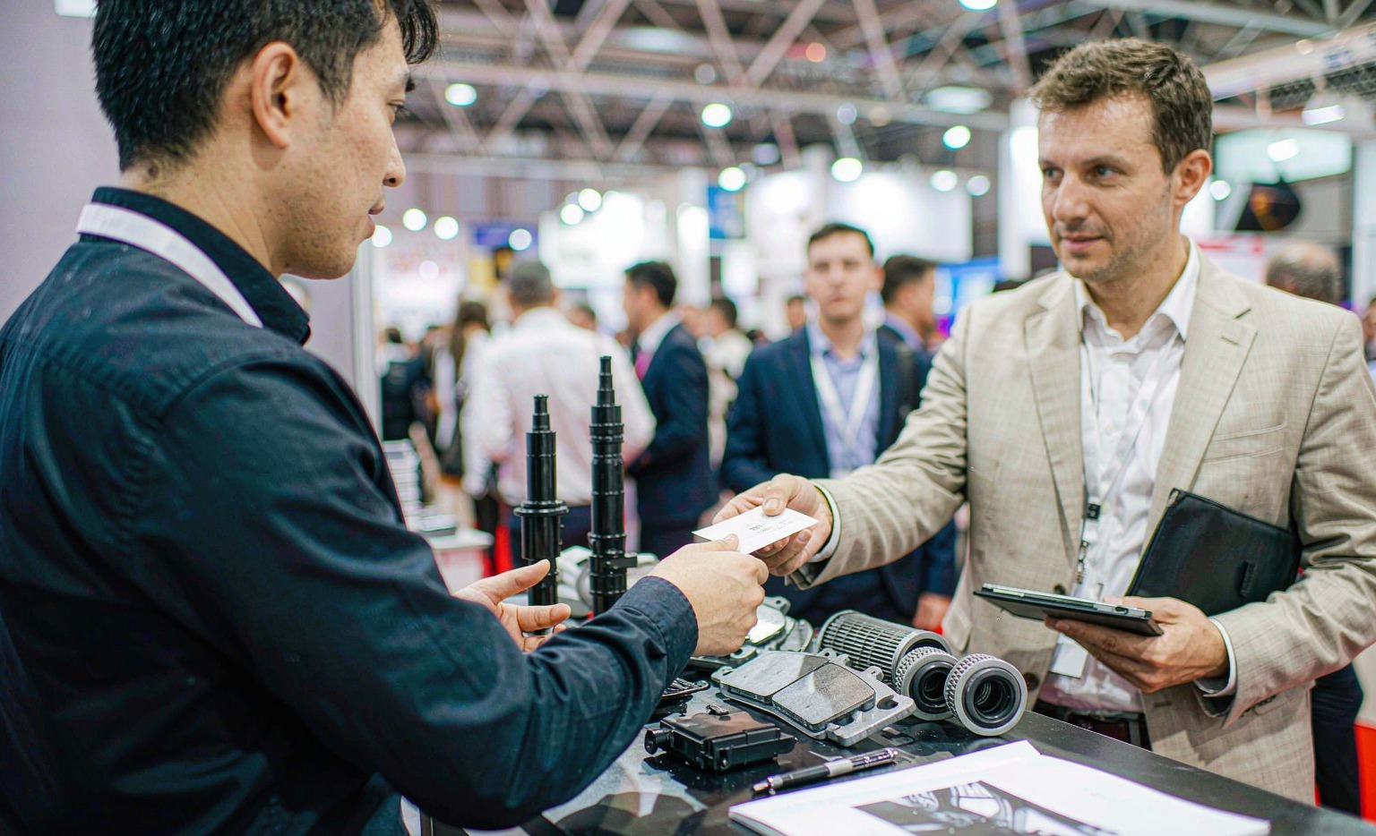 B2B trade fair booth interior showing a Chinese aftermarket auto parts supplier sample table with brake pads, oil filters, ignition coils and an ECU module, sales representative handing a sample to an overseas buyer