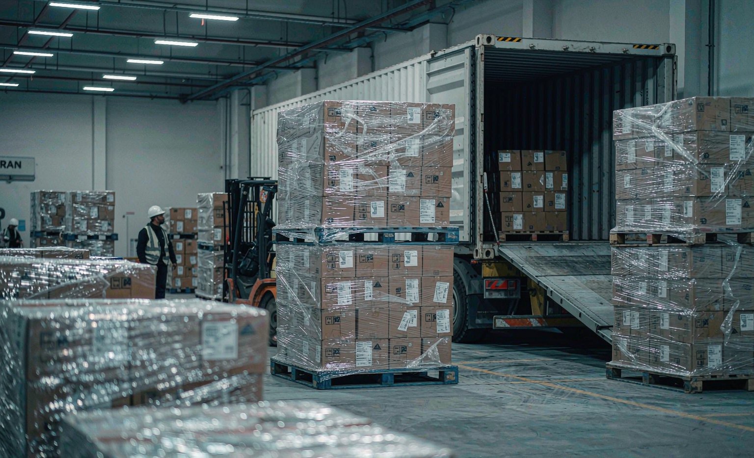 Export warehouse loading bay with pallets of aftermarket parts wrapped in clear stretch film and an open 40HQ shipping container with a forklift ramp leading up to it