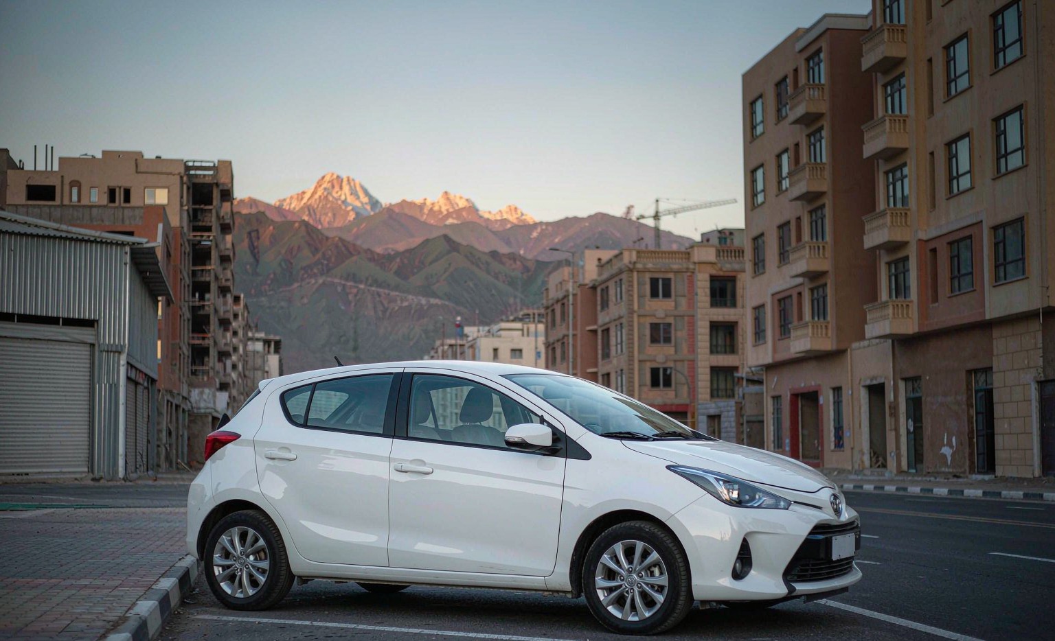 Tehran street in late afternoon with a clean modern Japanese compact sedan parked at the kerb — the visual anchor of the Iranian demand for Japan-built stock in 2026