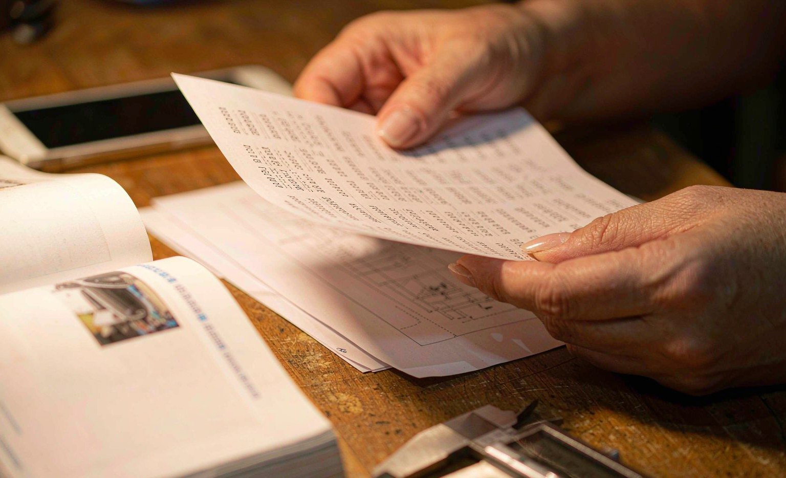 Close-up of hands holding a printed OE number list beside a parts catalogue and a smartphone showing a fitment search, representing OE number check and cross-reference verification for auto parts sourcing