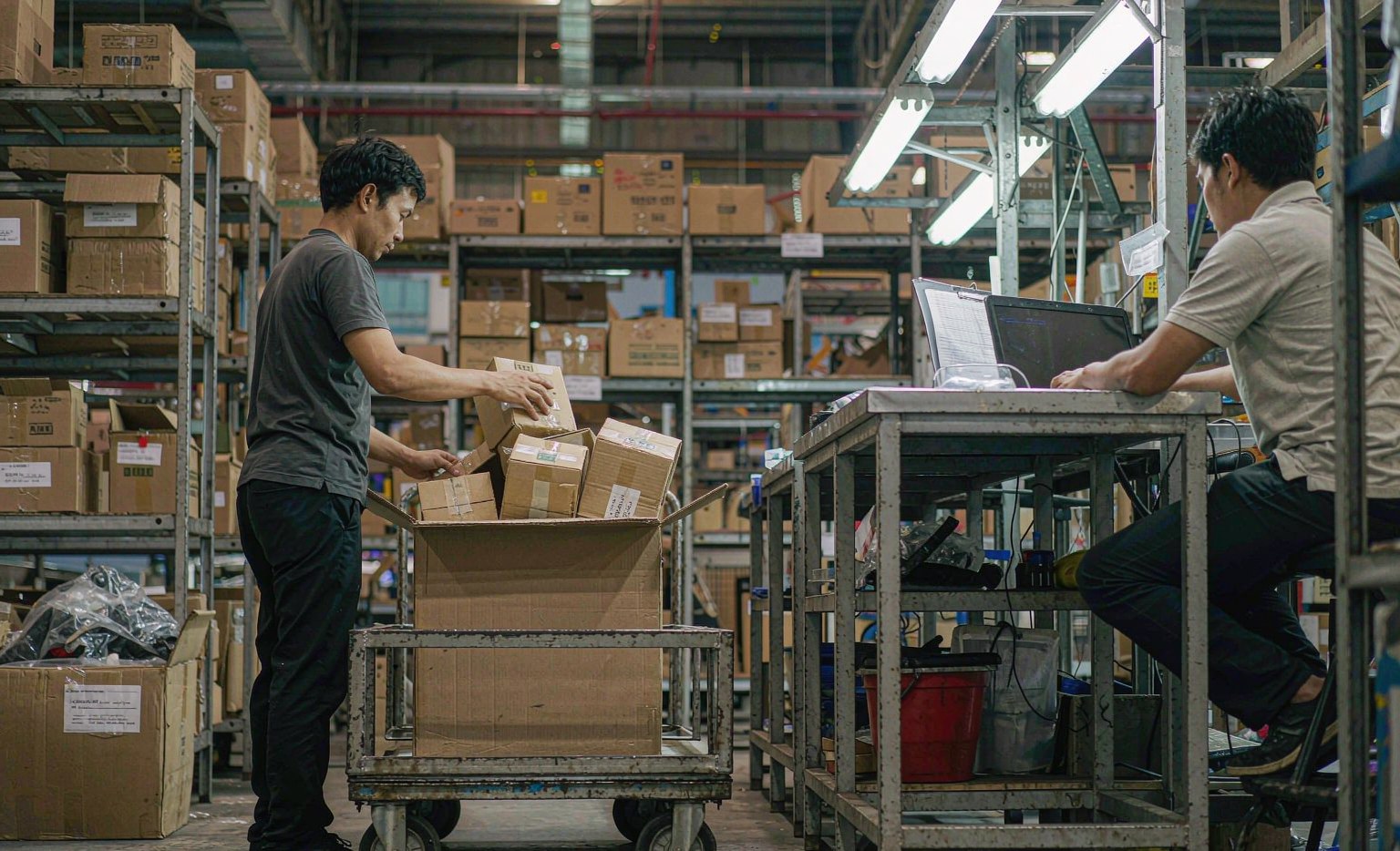 Warehouse worker packing boxed auto parts while a colleague checks an online order list on a laptop, representing year-round online auto parts sourcing from China after the Canton Fair